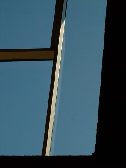 A view of a clear, bright blue sky through a window. The tan-colored wooden frame bisects the image, with a clear blue sky visible through the glass. The right edge reveals a dark, undefined mass of interior space creating strong contrast. Light falls sharply across the frame, defining its shape.