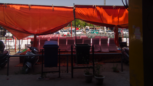 A photo of a stage for a political rally, with two trash cans in the foreground. In the foreground, there is a blue and a green trash can. In the middle distance, there is a stage set up with multiple chairs dressed in white. The stage has a canopywhich is saffron/orange in colour, and this casts a coloured glow on the chairs on the stage. In the background, there are audience chairs, some covered in white, others bare. In the far background, there are a bunch of parked cars.