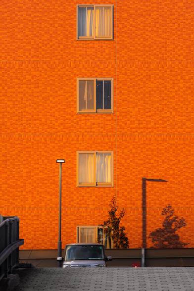 The image shows a vibrant orange building with a grid pattern. It has three windows, each with curtains. A car is parked below in front of the building, partially visible. There are shadows of a lamp post and a tree on the wall, indicating the direction of the light source. The setting appears to be a residential area, possibly late in the day with warm lighting.