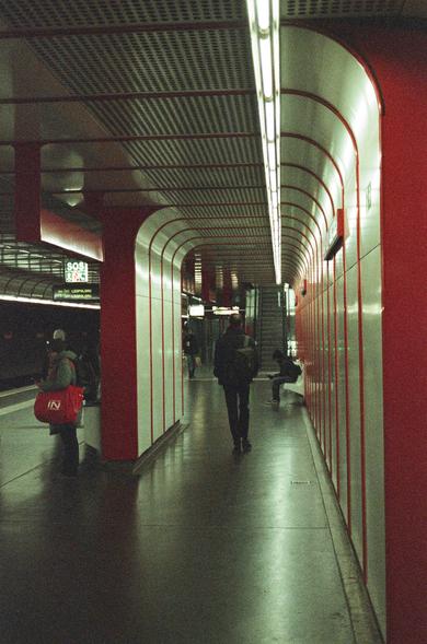 The image shows an underground metro platform. The station has a modern design with red and white wall panels and columns. There are people walking on the platform, and someone is sitting on a bench. The atmosphere suggests it's a regular day at the metro.
