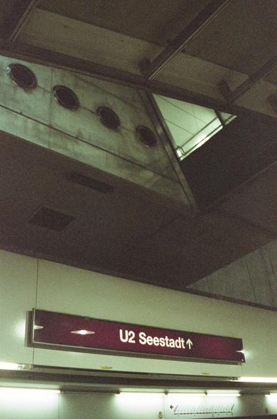 The image shows a subway or metro station with a sign indicating "U2 Seestadt." The sign suggests a directional arrow pointing upwards. The station's architecture is visible, including some ceiling vents and structural elements. The lighting is dim, giving a slightly industrial feel to the setting.