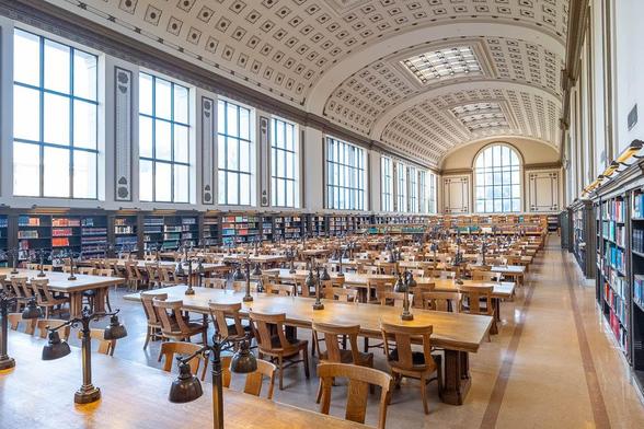 A grand, neoclassical, well-lit library with high arched ceilings and large windows. Rows of wooden tables with chairs and reading lamps fill the space. Shelves filled with books line the walls.