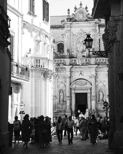 A monochrome scene of a city in Italy with an elegant historical church straight ahead, and the crowd silhouetted against it.