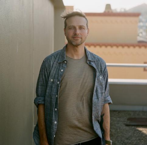 Billy Gomberg, wearing a blue chambray button down shirt and grey tee, gives a serene look directly in the camera as his hair is tousled by the afternoon sea breeze on a San Francisco rooftop