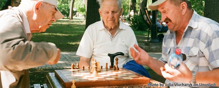 Two senior men playing chess with a third senior man looking on. They are in a park-like setting adjacent to a seniors community.