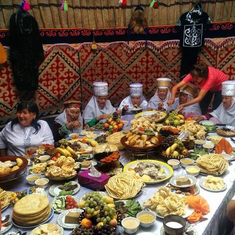 Foto dentro de una yurta kazaja. Una mesa grande llena de conmida, panes y frutas. Alrededor hay 8 mujeres. Las más mayores llevan un vestido tradicional kazajo con cofia. Están comiendo.