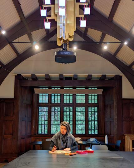 Artist Jenie Gao carving a circular woodblock at a large table in a heritage building with a barrel ceiling and a large bay window. The building is constructed with old dark wood paneling. There is a modern looking chandelier made up of geometric shapes hanging from the ceiling, in contrast with the surroundings.

Jenie is a person of Taiwanese-Chinese descent. They have salt-and-pepper black hair and in this photo are wearing layered black and gray sweaters and a scarf.