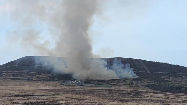 A photo of the same fire taken about 20 minutes later from further away. It has spread across the hillside and there is a thick plume of grey smoke rising into the blue sky.