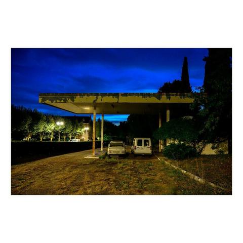 Une ancienne station-service abandonnée, dont le toit en métal est écaillé, se dresse sous un ciel bleu nuit profond. Deux véhicules vieillissants, une voiture beige et une fourgonnette blanche, sont garés sous l’auvent. La végétation commence à envahir les abords de la station, avec des herbes hautes et des buissons grimpants.