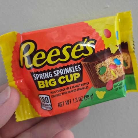 Photograph of a Reese's Spring Sprinkles Big Cup held by a white person's hand against a light colored wall. The packaging has the name of the candy and the Reese's logo. On the right there is a cut of a Reese's Peanut Butter Cup with multi-colored sprinkles inside of it.