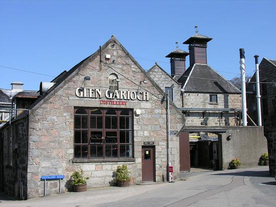 Glen Garioch Distillery. The image shows a stone distillery building with its gable turned towards the camera in the left foreground. Stills can be glimpsed through a large window. Further right and further away are more buildings, topped off by two square slate roofs with pagodas. The sky is blue and the distillery in bright sunlight.