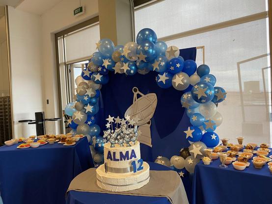 A picture of a celebratory cake with white frosting on a small table, with 12 ALMA-like antennas on top, together with 12 stars, and ALMA and 12 written in bold blue letters.

Behind, a larger table has a round cloth with a white ALMA-like antenna printed on it, surrounded by white, light blue, and dark blue balloons with stars. That cloth is surrounded by additional things to eat, like plates of fruit, parfaits, jellies, and other delicacies.