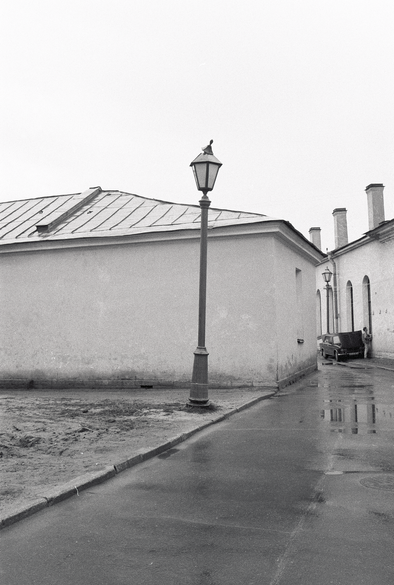 A narrow lane between featureless buildings.  A lamppost.  In the distance a single car nestled in the alley with the hood open.
A figure stands by the car.  Black and white photo from 1991, somewhere in Russia.