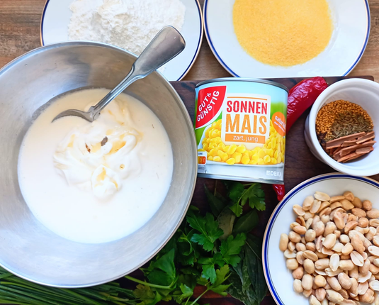 Auf dem Tisch eine Dose Mais, auf Tellern Polenta, Mehl eine rote Chili, Petersilie + Schnittlauch, eine Schale mit Erdnüssen, eine Schüssel mit Frischkäse + Ahornsirup + eine Schale mit den Gewürzen