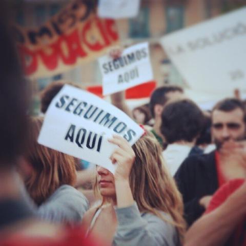 A girl, in the middle of the people, shows in her hand a banner with the message "Still here".