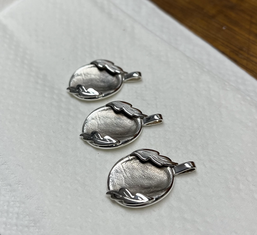 Three silver pendants in a row on top of a white paper towel. Each pendant is a fingerprint indent with two oak leaves surrounding it.