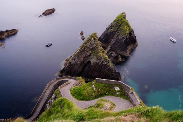A scenic view of Dunquin Pier in County Kerry, Ireland, featuring winding stone paths leading down to the sea, triangular rocky outcrops covered with green grass, and small boats floating in calm waters.