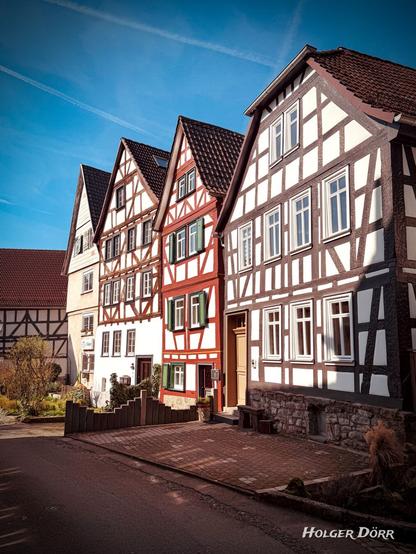 Eine malerische Fachwerkhauszeile in Laubach, Hessen, bei strahlend blauem Himmel. Die historischen Gebäude sind in verschiedenen Farbkombinationen gestaltet, darunter ein auffälliges rot-weißes Fachwerkhaus mit grünen Fensterläden. Sonnenlicht betont die Strukturen der Häuser und verleiht der Szene eine warme, einladende Atmosphäre. Im Vordergrund ein gepflasterter Eingangsbereich mit einer kleinen Bank und etwas Grün. Die Idylle spiegelt den traditionellen Charme der hessischen Fachwerkstädte wider.