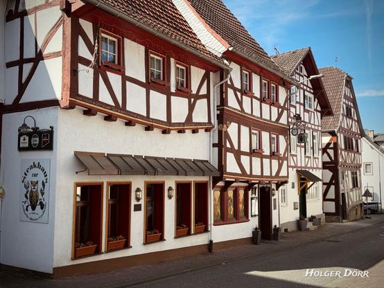 Ein idyllisches Straßenbild in Laubach mit historischen Fachwerkhäusern unter blauem Himmel. Im Vordergrund befindet sich das „Steakhaus zur Eule“, ein traditionelles Gasthaus mit weißer Fassade, dunklem Fachwerk und roten Fensterläden. Über dem Eingang hängt ein Wirtshausschild mit Biermarken, und Blumenkästen schmücken die Fenster. Die angrenzenden Gebäude setzen die malerische Fachwerkarchitektur fort, mit filigranen Holzdetails und kleinen Fenstern. Die Straße wirkt ruhig und einladend, das Sonnenlicht wirft weiche Schatten auf die Fassaden
