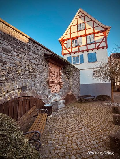 Ein malerischer, gepflasterter Platz in Laubach mit dem historischen Grünemannbrunnen. Der Brunnen ist in eine alte Steinmauer eingelassen, mit einem kunstvollen Sandsteinrelief verziert und hat einen steinernen Trog darunter. Davor stehen schmiedeeiserne Bänke mit Holzlatten, die zum Verweilen einladen. Im Hintergrund ragt ein auffälliges Fachwerkhaus mit roten Balken und blauen Fensterläden über die Mauer hinaus. Die warme Abendsonne taucht die Szenerie in ein goldenes Licht, während der Himmel in klarem Blau erstrah