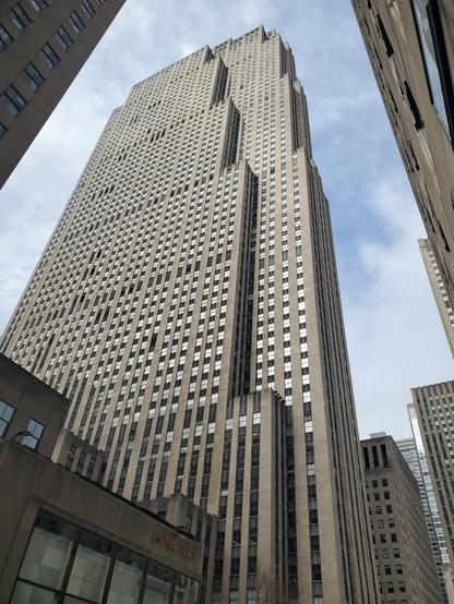 A view of 30 Rockefeller Plaza on a cloudy day. The photo is taken from a vantage point near the building's base, less than a block away from the southeastern corner, so the view is necessarily somewhat foreshortened. The tower rises high into the sky, with Art Deco detailing in vertical-line motifs that accentuate the building's height and the perspective view. It also has some staircase-like setbacks as it climbs.