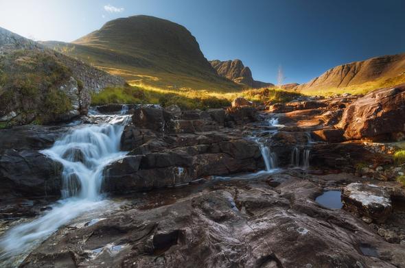 A serene highland scene showing a small, multi-tiered waterfall cascading over dark, rocky steps in bright sunlight, with a steep, grass-covered mountain rising behind it and a clear blue sky above, creating a striking contrast between the rushing white water in the foreground and the golden-lit slopes of the Applecross Peninsula in the distance.
