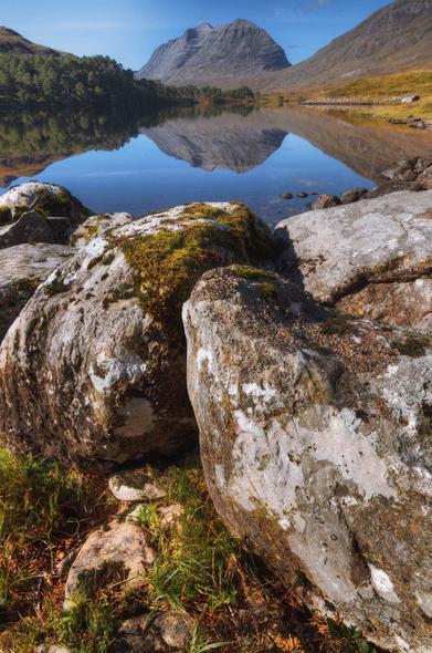 A tranquil Highland scene with large, weathered boulders in the foreground bearing patches of moss and lichen, leading to the mirror-like surface of Loch Clair reflecting the imposing peak of Liathach rising beneath a bright blue sky, with the surrounding slopes tinged in muted autumnal shades that accentuate the stillness of the water.
