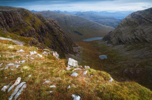 A dramatic mountain vista captured from a high ridge, with steep cliffs plunging into a broad, winding valley below where two small lochans nestle among golden-brown moorland and rugged slopes, all framed by a hazy, layered backdrop of distant peaks under a subdued, overcast sky.
