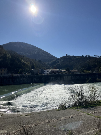 Foto eines Flusses der mit weißem Wasser über ein Wehr fällt . Im Hintergrund blauer Himmel und bewaldete Hügel. Auf einem ist ein Turm.