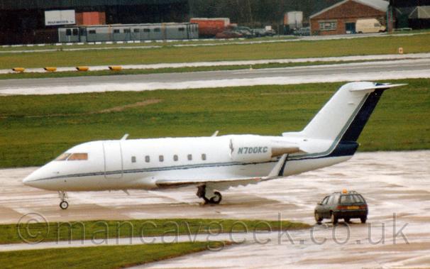 Slightly blurry side view of a white, twin engined BizJet taxiing from right to left. 
There is a thin blue stripe running along the body,. sweeping up to fill the rear of the tail.
The same blue stripe fills the rear of the up-turned wingtips.
The registration "N700KC" is on the side of the engine pods mounted on the sides of the rear fuselage.
A grassed area and concrete apron fill the foreground, with more grass and a runway leading up to black hangars in the distance.
