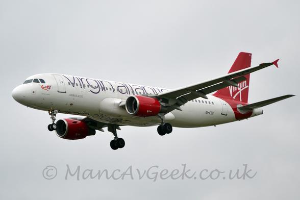 Side view of a white, twin engined jet airliner flying from right to left at a low altitude, with undercarriage lowered and flaps extended from the rear of the wing, suggesting it is about to land.
The plane has purple "Virgin Atlantic" titles running along the body, repeated along the planes underside,  with red engine pods slung under the wings, and red rear fuselage and tail.
There is seemingly hand-written white "Virgin" titles on the tail.
Grey cloud fills the rest of the frame.