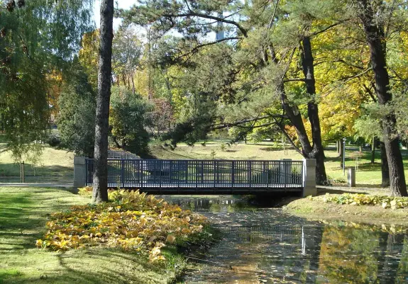The image depicts a serene park scene featuring a bridge over a small waterway or stream. Surrounding the bridge are lush green trees and colorful autumn foliage, suggesting the season is fall. There are manicured lawns and fallen leaves along the banks of the water, contributing to the tranquil atmosphere of the setting.

Image Credits: Wikimedia / Frees / CC0