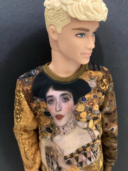 Three-quarter slightly downward angle picture of the head and upper body of a blonde Ken doll — the prince from Fairytopia — wearing a long-sleeved shirt with a vivid reproduction of Portrait of Adele Bloch-Bauer by Gustav Klimt. The design covers the entire chest of the garment. The doll’s gaze is fixed on the lower right of the frame.