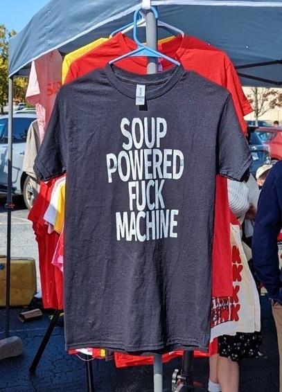 A black t-shirt displayed on a hanger at an outdoor market. The shirt has bold white text that reads:

“SOUP POWERED FUCK MACHINE.”