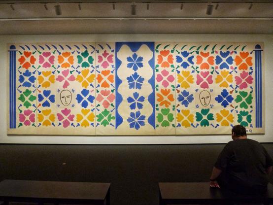 A man sits on a bench in a dimly lit gallery, looking at a large, very long work by Matisse featuring colorful, flower-shaped cutouts, with two drawings of heads.