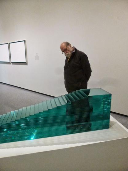 A man in dark clothing with white beard and glasses looks at the sculpture, a shaped stack of green-colored panes of glass.