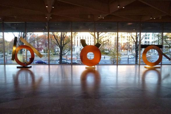 The main element in the gallery is three large sculptures of circles of metal, placed in front of a wide window showing part of a city street, the sculptures reflected in the shiny, marble floor.