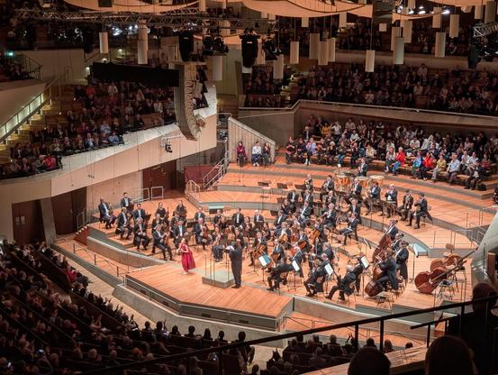 Das Bild zeigt eine klassische Konzertveranstaltung in einem modernen Konzertsaal. Im Zentrum des Bildes befindet sich eine Orchesterbühne, auf der eine Orchestergruppe in formeller Kleidung sitzt und spielt. Die Musiker sind in einer traditionellen Konzertformation angeordnet, mit Streichinstrumenten im Vordergrund, Blasinstrumenten in der Mitte und Schlagzeuginstrumenten im Hintergrund. Vor der Bühne steht ein Dirigent, der die Musiker leitet, und eine Violinistin in einem roten Kleid, die sich auf der Bühne befindet.

Der Konzertsaal ist gut besucht, mit Zuschauern auf mehreren Ebenen, die das Geschehen beobachten. Die Architektur des Saals ist modern, mit hohen Decken und einer offenen Struktur, die den Blick auf die Bühne freigibt. Die Beleuchtung ist warm und konzentriert sich auf die Bühne, während der Rest des Saals in etwas dunklerem Licht gehalten ist. Die Atmosphäre wirkt formell und kulturell, typisch für klassische Konzerte.