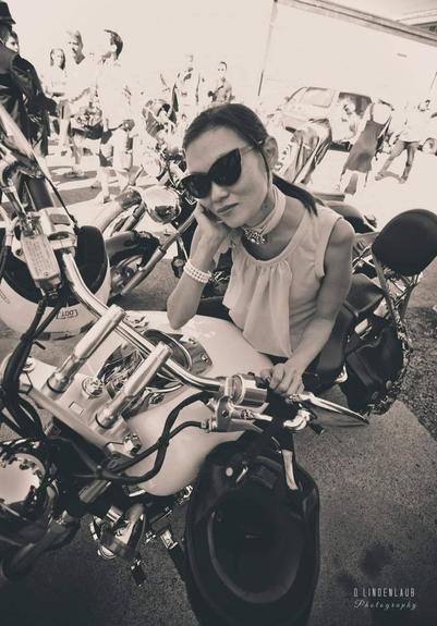 Monochrome shot of me, positioned prominently on a white motorcycle. Handlebars are chrome, detailed. Soft focus on the background showing pedestrians and parked vehicles. High contrast and desaturation creates a vintage aesthetic. "D. Lindenlaub Photography" watermark in the bottom right corner.