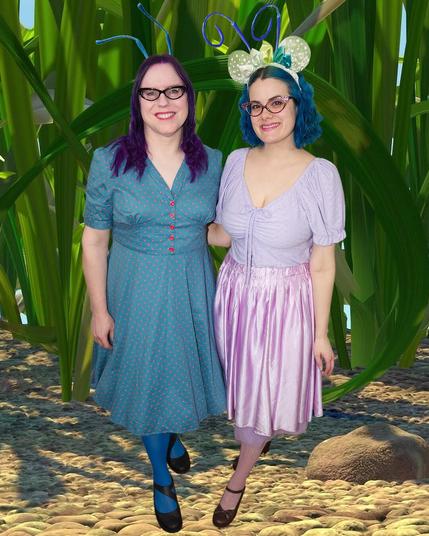 The image features two women standing side-by-side, posing for the camera against a backdrop of tall green plants.

Both women have long dark hair and are dressed in vintage-inspired attire. The woman on the left wears a blue dress with short sleeves and buttons down the front, complemented by blue tights and black shoes. Her counterpart on the right sports a lilac dress with short sleeves and a lace top, paired with brown heels.

The women stand on a sandy surface, surrounded by large green plants that stretch towards the sky behind them. The overall atmosphere of the image suggests a sunny day at the beach or in a tropical location.