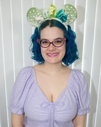 The image features a woman with vibrant blue hair styled in two ponytails, wearing a light purple dress adorned with small white dots. She sports a pair of mouse ears headband on her forehead, adding a playful touch to her overall appearance.

In the background, a wall composed of vertical blinds provides a subtle yet functional setting for the subject. The woman's attire and accessories suggest she may be attending an event or celebration, possibly related to Disney or another theme that incorporates mouse ears as part of its branding. Overall, the image presents a cheerful and lively atmosphere, capturing the essence of a fun-filled occasion.