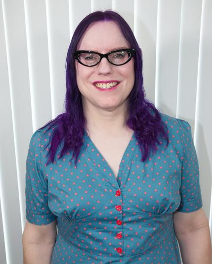 The image features a woman with long, dark purple hair that falls just below her shoulders. She is wearing a teal blue top adorned with small red polka dots and has three red buttons down the front. The woman's attire is complemented by black-framed glasses and a subtle smile, as she gazes directly at the camera.

In the background, white vertical blinds are visible, providing a clean and minimalist setting for the subject. Overall, the image presents a straightforward yet visually appealing composition that showcases the woman's unique style and personality.