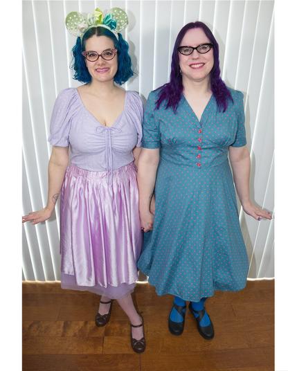 The image features two women standing side-by-side against a white wall adorned with vertical stripes. The woman on the left has dark blue hair styled in curls, complemented by a purple dress featuring short sleeves and a V-neckline. Her counterpart on the right boasts long, straight purple locks, paired with a teal dress boasting a V-neckline and short sleeves.

Both women wear black shoes, while the woman on the left sports a bracelet on her right wrist. The background of the image is characterized by a white wall with vertical stripes, providing a clean and minimalist backdrop for the subjects.