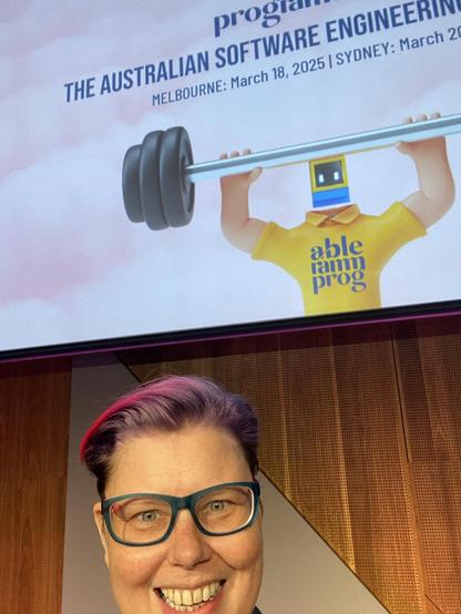 A selfie of me smiling before my Melbourne locknote, I have pale skin and purple and pink short hair and turquoise and pink glasses. There is a massive screen behind me with the programmable mascot lifting a barbell.