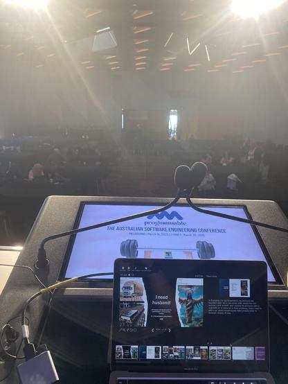 The speakers view in Melbourne. Visible is the lectern, my laptop with my creepy AI slop introduction slides, a confidence monitor showing the hold screen, and bright lights. Beyond the bright lights there are silhouettes of the audience.