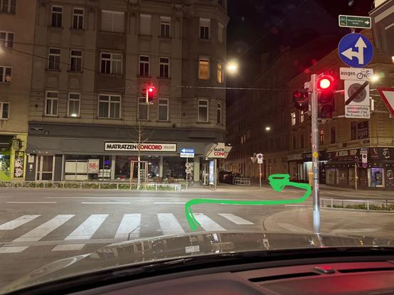 A nighttime street scene showing a traffic light with a red signal and a road signs shows direction straight and right. There is green arrow marking a turning lane right and then left between the buildings.