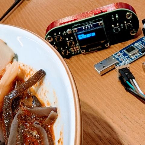 Bowl of ramen in foreground, small game console and device for uploading firmware behind it.