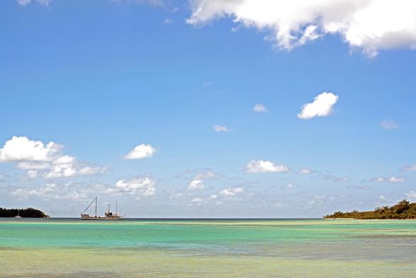 Photo shows a freighter or fishing boat on the horizon line where the green yellow blue pacific ocean meets a clear blue sunny sky with some white clouds. 2/3 of the photo is sky to show dimension. On each side of the boat there is small inlet of land bordering the natural harbour the boat is entering. It is a sunny, peaceful day on an island in the South Pacific.