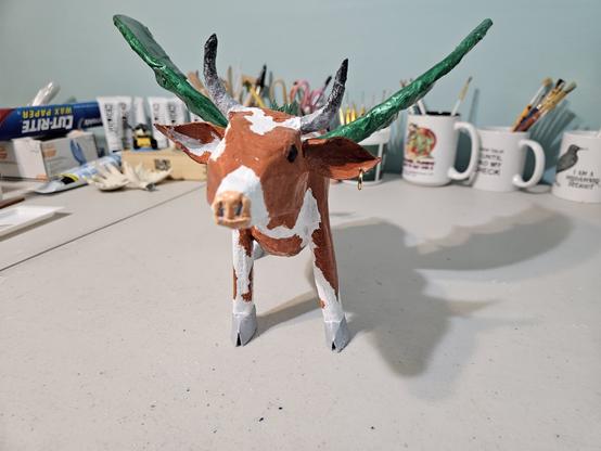 Guernsey cow-dragon, facing the camera.  It has a tilt to its head, as if it is challenging the observer to a stare down.  Or worse.  The cow has green, leathery bat wings, and a brass ring in its ear.