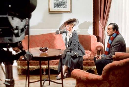 Thulin (wearing a grey suit and large hat) and Visconi sit on classic, old couches and look in the distance. A camera is seen on the left. The set is lit.